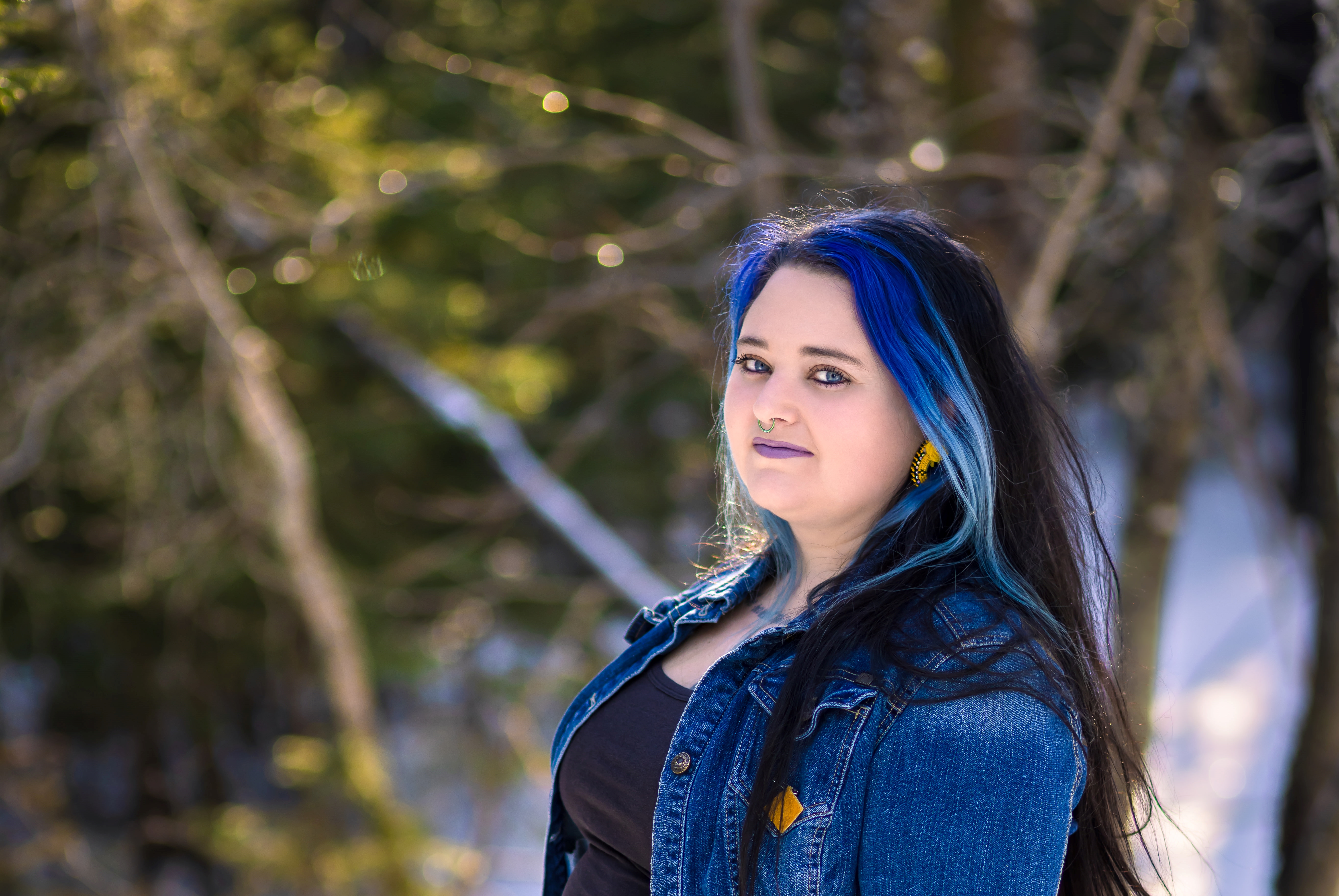 A young woman stands among the trees. She has long flowing black hair with blue highlights around her face.
