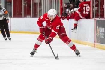A young man is on the ice with his stick down poised for a shot. He wears a red hockey uniform.