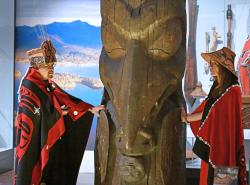 A man and a women dressed in button blankets and cedar hats stand with their hands on the totem pole.