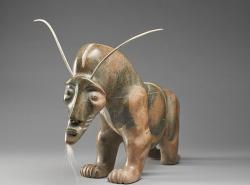 A mottled brown soapstone carving shows a muscular Muskox with an exaggerated animated face with a tuft of white fur coming out of his chin and narrow horns sprouting from the top of its head. 