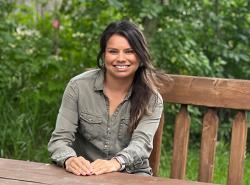 A young woman sits at on a bench behind a wooden table. It's outdoors and there is a leafy backdrop. She has her hands folded on the table in front her. She is smiling widely.