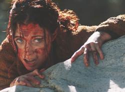 A woman with a bloodied face clings to a rock cliff.