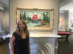 A woman stands in from of a framed painted landscape. Hanging in the middle of the painting is a beaded medallion that says "Land Back"