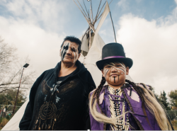 A woman and boy stand outdoors in front of a tipi. Their faces are painted with black and white paint. He wears a top hot with a purple ribbon wrapped around it. He has on a purple vest with two white feathers appliqued on the shoulders.