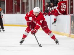 A young man is on the ice with his stick down poised for a shot. He wears a red hockey uniform.