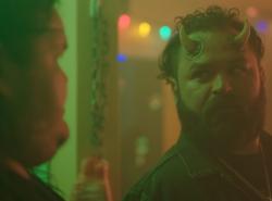 A bearded man with horns growing from his forehead is seen with another man in a room with Christmas decorations hanging, including multi-coloured strings of lights.