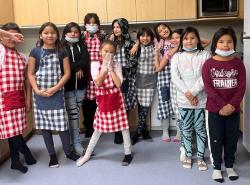 A group of children, mostly girls, dress in red or blue checked aprons in a bright kitchen.