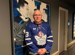 A man in a Toronto Maple Leafs jersey stands beside a large drawing featuring former Toronto Maple Leafs captain George Armstrong, whose mother was from the Kitigan Zibi Nation.