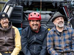Three men sit on the tailgate of a vehicle laughing togeter. They are wear hats of some kind, but the man in the middle wears a red bike helmet.