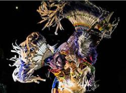 A fancy dancer is photographed in the air in a spotlight against a black background